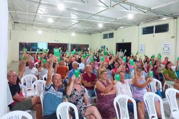 Um grande grupo de associados participa de uma assembleia da Sicredi Caminho das Águas. Sentados em cadeiras brancas, levantam cartões verdes com a palavra "Assembleia", demonstrando engajamento e participação no evento cooperativo.