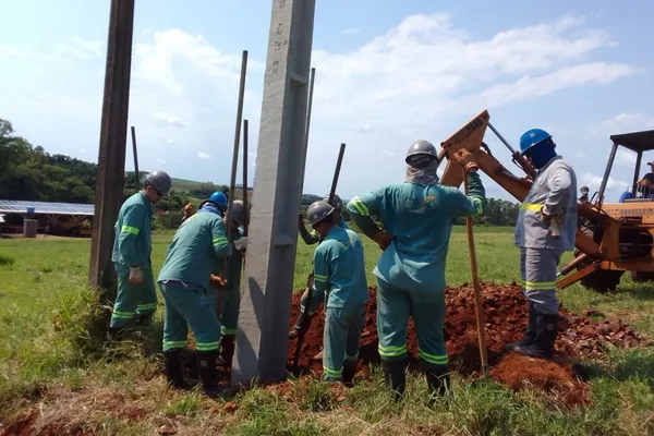 Equipe de trabalhadores da Cermissões instala um poste de concreto em uma área rural sob forte calor. Eles utilizam uniformes de segurança e ferramentas manuais, enquanto uma retroescavadeira auxilia no processo. O céu está parcialmente nublado.