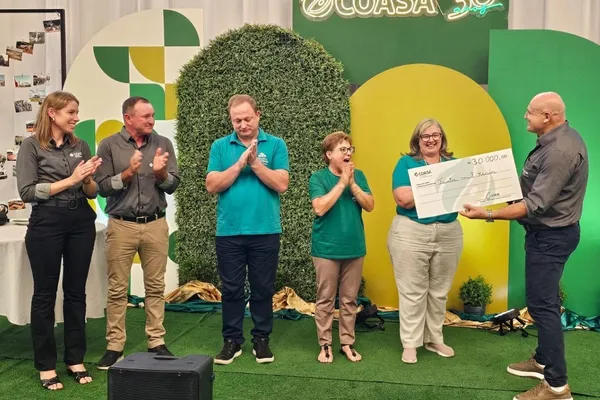 Seis pessoas participam de uma cerimônia de entrega de doação da Coasa. Um homem segura um cheque simbólico de R$ 30 mil, enquanto os demais aplaudem e demonstram alegria. O cenário conta com decoração verde e amarela, incluindo um painel com o logotipo da Coasa.