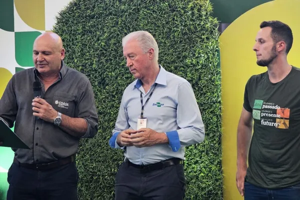 Três homens participam de um evento da Coasa e Embrapa. Um deles, usando camisa da Coasa, fala ao microfone segurando um documento. Outro, com camisa da Embrapa, observa atentamente, enquanto o terceiro, com camiseta verde, acompanha a apresentação.