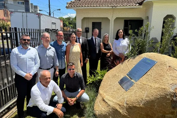  Um grupo de nove pessoas posa para uma foto ao ar livre em frente a um prédio de arquitetura tradicional, com telhado de cerâmica e paredes claras. Eles estão ao lado de uma grande pedra com uma placa de metal fixada, que contém informações sobre o lançamento da pedra fundamental do memorial da Sicredi Caminho das Águas. O ambiente conta com vegetação ao redor da pedra e céu azul ao fundo, indicando um dia ensolarado. Alguns dos participantes estão de pé, enquanto outros estão agachados, todos sorrindo para a câmera.
