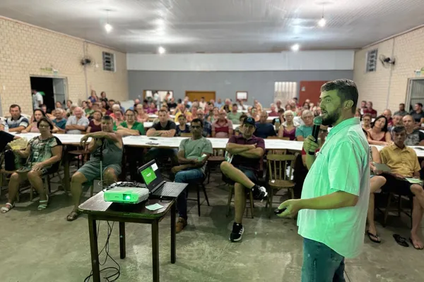 A imagem mostra um homem de camisa clara e barba segurando um microfone enquanto fala para uma grande plateia em um salão comunitário. O público, composto por homens e mulheres de diferentes idades, está sentado em mesas longas, demonstrando atenção à apresentação. Na frente, há um projetor e um notebook exibindo um slide com fundo verde. O ambiente tem paredes de tijolos e iluminação simples, sugerindo uma reunião de caráter informativo, possivelmente ligada a uma cooperativa ou associação local.