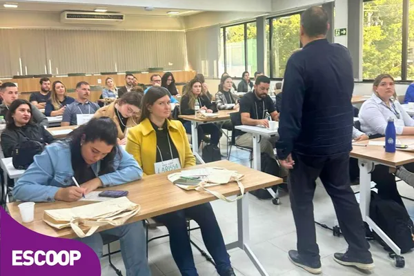 Sala de aula com participantes atentos durante atividade formativa da Escoop; destaque para crachás e materiais sobre mesas.