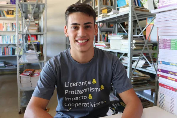 Jovem sentado em biblioteca escolar, usando camiseta com os dizeres "Liderança & Protagonismo & Impacto & Cooperativismo", sorri para a câmera em meio a prateleiras com livros e pilhas de apostilas.