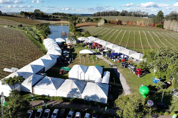 Vista aérea de evento com tendas brancas entre vinhedos, máquinas agrícolas e estandes, ao lado de lago em área rural.