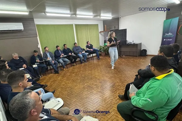 Participantes da Cotrijuc assistem a uma apresentação em formato de roda durante a abertura da Academia de Líderes, com apoio da Escoop. A palestrante está de pé, interagindo com o grupo. Banners do Sistema Ocergs e Escoop estão ao fundo.