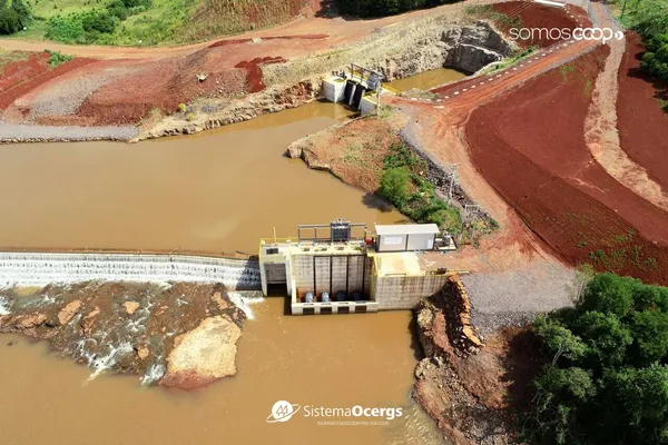 Vista aérea da PCH Linha Onze, da Ceriluz, em Coronel Barros, com barragem, vertedouro e estruturas de geração de energia integradas à paisagem natural e ao solo avermelhado da região. 
