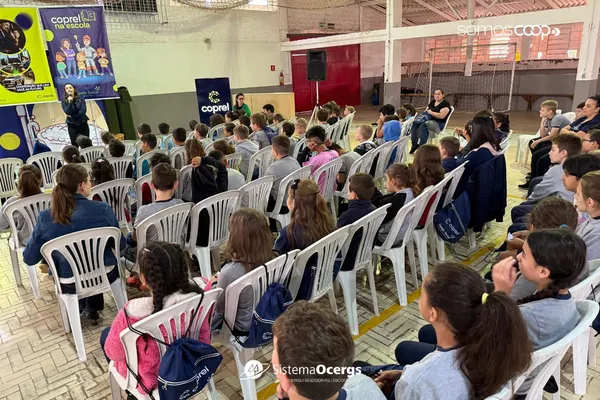 Crianças participam atentas do projeto Coprel na Escola, em Santa Cecília do Sul, sentadas em cadeiras brancas, enquanto um palestrante conduz a atividade ao lado de banners da Coprel. 
