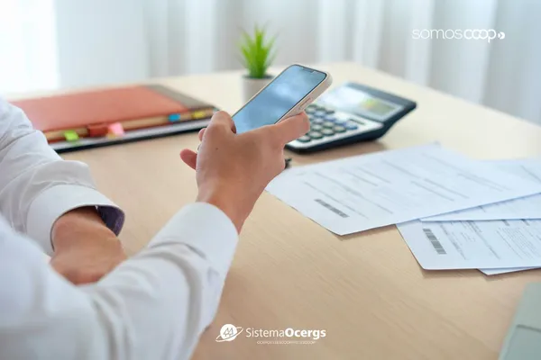 Pessoa segura um celular enquanto confere contas e boletos sobre uma mesa, ao lado de uma calculadora e materiais de escritório. A cena representa o uso da fatura digital para facilitar o controle financeiro, destacando praticidade e tecnologia no cotidiano.