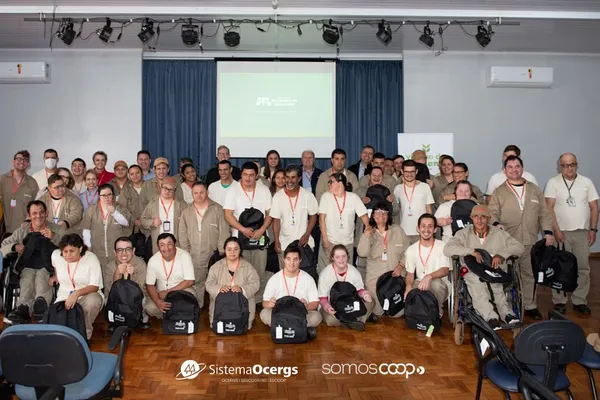 Integrantes do projeto Viveiro de Cidadania, da Cotrijal, posam em grupo durante o lançamento do Programa de Capacitação “Sementes de Cidadania”. Eles vestem uniformes e seguram mochilas do evento, com destaque para a diversidade e inclusão. Ao fundo, aparecem os logos do Sistema Ocergs e do movimento SomosCoop.