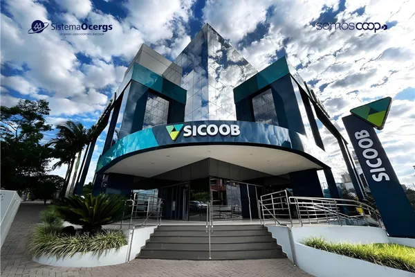 Fachada de uma agência do Sicoob, com arquitetura moderna em vidro espelhado, céu azul com nuvens e jardim na entrada.