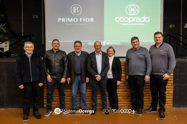 Lideranças da Cooprado e autoridades posam sorrindo em frente à projeção das novas marcas da Cooprado e Vinhos Primo Fior, durante evento em Antônio Prado.