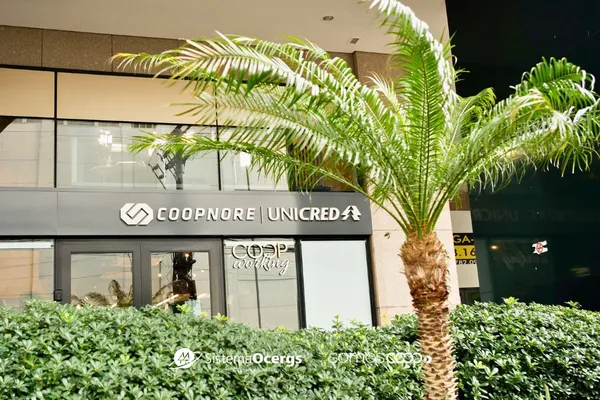 Descrição da imagem: Fachada da sede da Coopnore Unicred, com destaque para o letreiro da cooperativa e a inscrição "COOPworking" em uma das portas de vidro. Em primeiro plano, há uma palmeira e arbustos que compõem a paisagem externa.