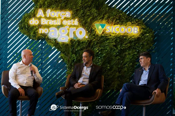 Francisco Reposse Junior, Miguel Oliveira e Marco Aurélio Almada participam de evento do Sicoob em frente a painel com a frase "A força do Brasil está no agro".