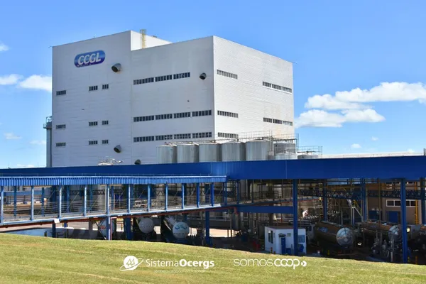 Fachada da indústria da CCGL em Cruz Alta (RS), com estrutura moderna, tanques de armazenagem e caminhões de transporte posicionados abaixo de passarelas azuis, sob céu limpo e gramado bem cuidado em primeiro plano.