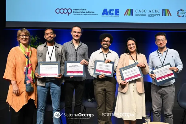 Participantes do evento da Aliança Cooperativa Internacional (ACI) posam sorridentes no palco com certificados de premiação, representando jovens pesquisadores reconhecidos por suas contribuições ao cooperativismo.