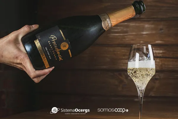 Pessoa serve espumante Aurora Procedências em taça personalizada; rótulo destaca-se com medalha dourada. Logos do Sistema Ocergs e SomosCoop aparecem na base da imagem.