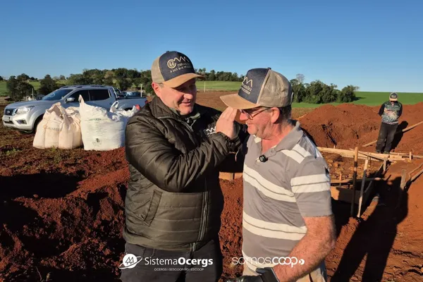 Dois homens, um deles vestindo jaqueta da Cermissões, sorriem e se cumprimentam em meio a uma obra no campo, com sacos de insumos, veículos e trabalhadores ao fundo. Céu limpo e solo avermelhado completam a cena.