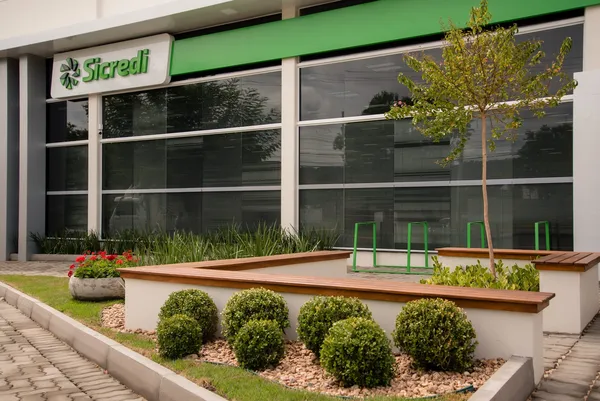 A imagem mostra a fachada de uma agência do Sicredi, com o logotipo em destaque, área externa ajardinada com arbustos, bancos de madeira e uma pequena árvore. O design é moderno, organizado e acolhedor.