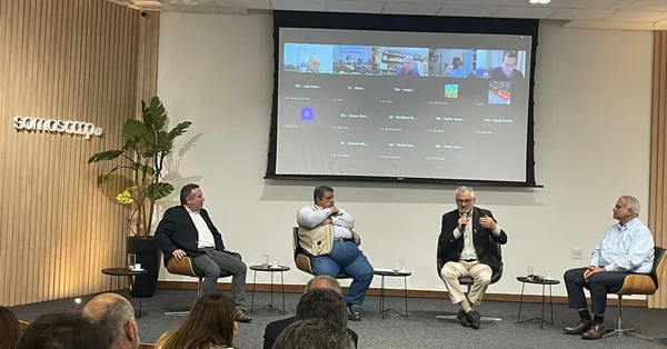 Quatro homens sentados em cadeiras em um auditório durante um painel de discussão, com um telão ao fundo exibindo participantes conectados virtualmente.