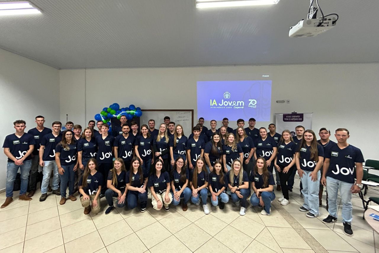 Grupo de jovens cooperativistas posa durante evento do programa IA Jovem da Coopermil, com camisetas personalizadas e projeção ao fundo.