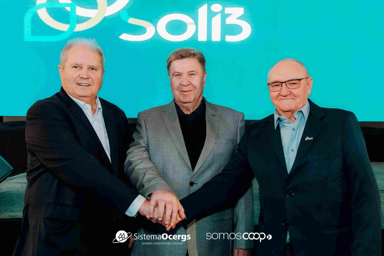 Mãos despejando grãos de soja em um monte, com céu azul ao fundo. A imagem transmite a força do agronegócio cooperativista, destacando a marca do Sistema Ocergs e o carimbo SomosCoop. 