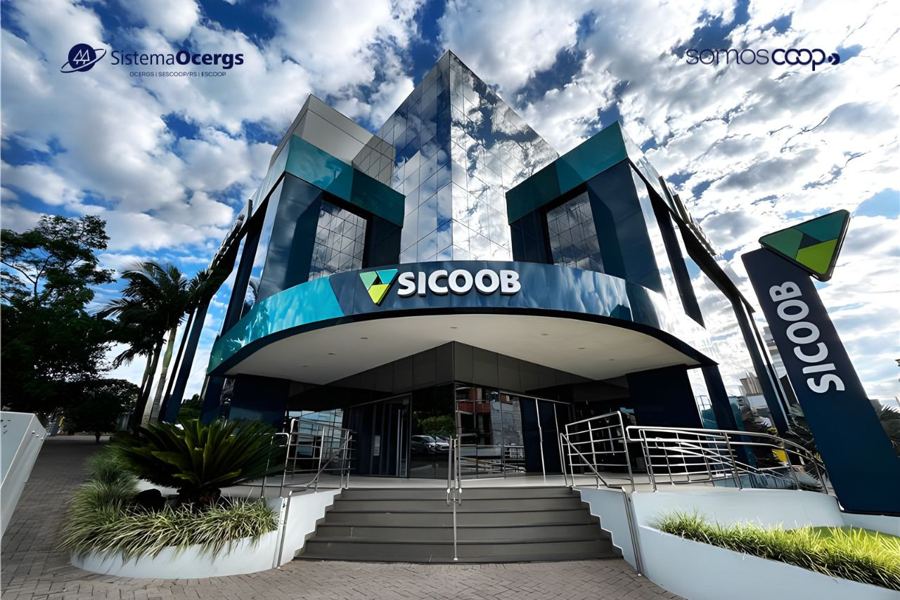Fachada moderna de uma agência do Sicoob com arquitetura espelhada, céu azul com nuvens ao fundo e logotipos do Sistema Ocergs e SomosCoop visíveis no topo da imagem.