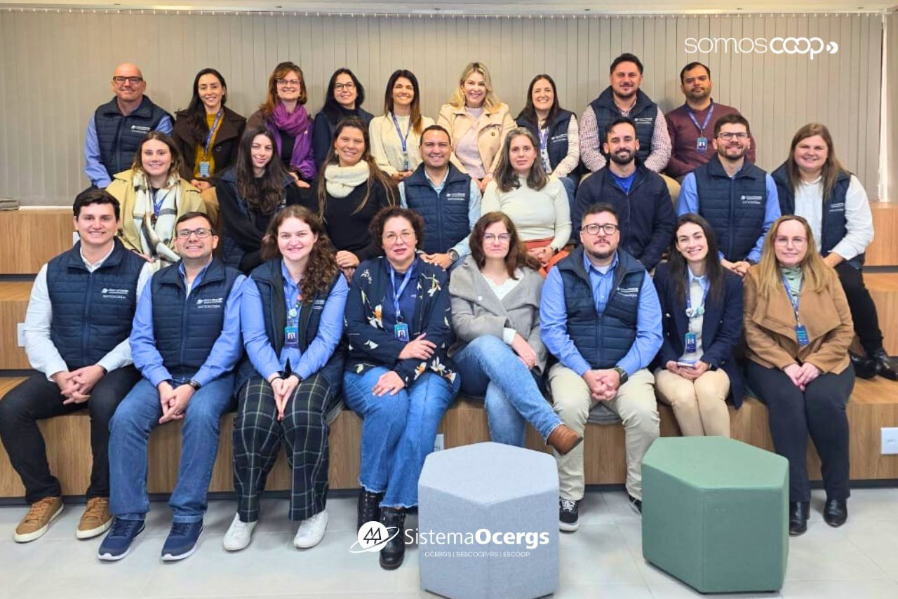 Equipe do Time de Soluções do Sistema Ocergs reunida em ambiente interno, posando para foto em arquibancada de madeira. O grupo, formado por homens e mulheres, veste coletes personalizados da instituição e demonstra clima de integração e profissionalismo.