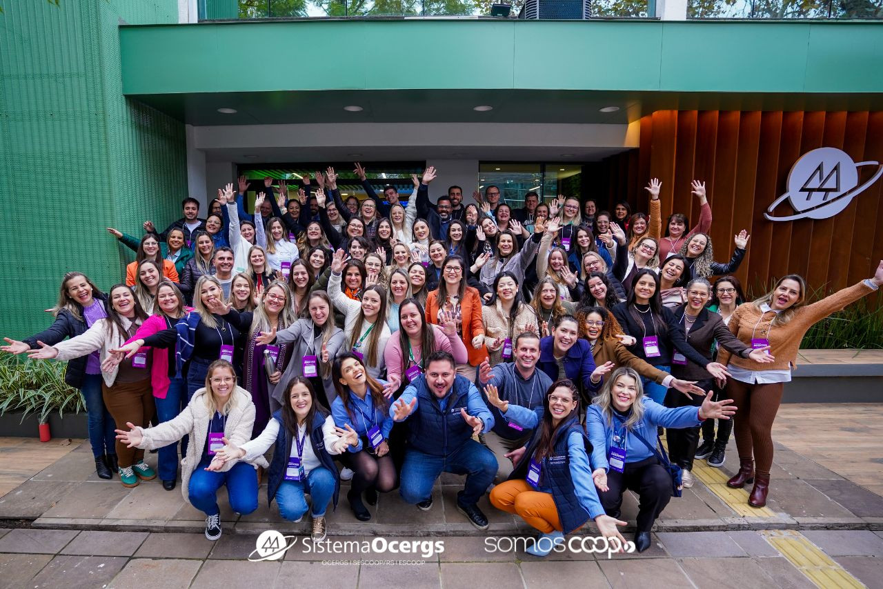 Grupo animado com dezenas de pessoas cooperativistas posa em frente à sede do Sistema Ocergs durante o Encontro de Agentes 2025, com gestos de celebração e integração.