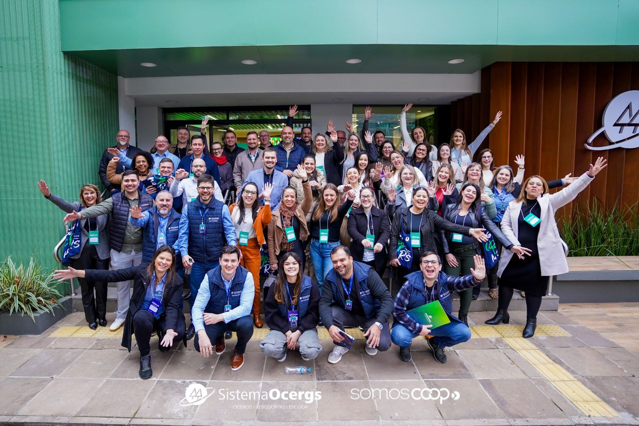 Grupo animado com dezenas de pessoas cooperativistas posa em frente à sede do Sistema Ocergs durante o Encontro de Agentes 2025, com gestos de celebração e integração.