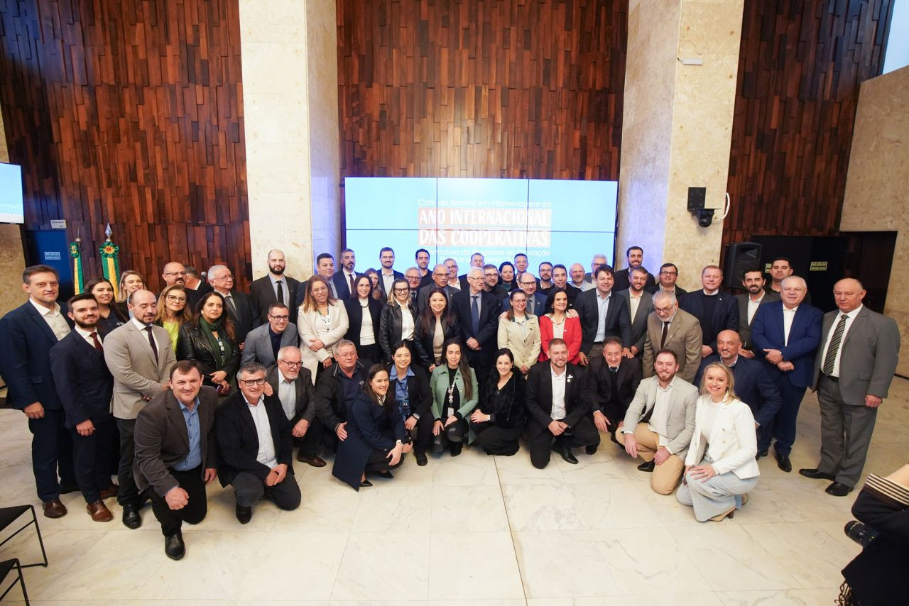 Grupo de dezenas de pessoas, entre homens e mulheres, posa para foto oficial em ambiente interno, diante de painel com os dizeres