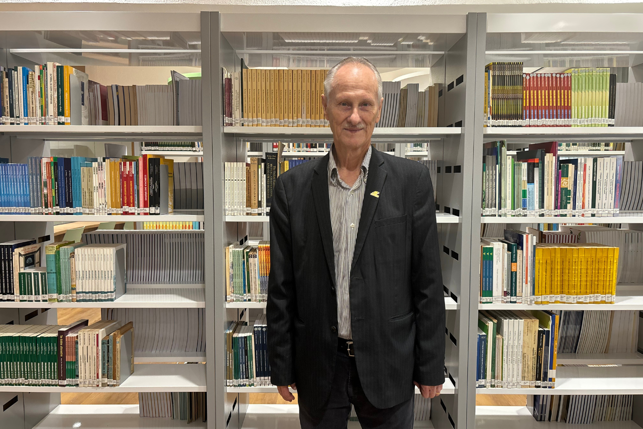 A imagem mostra Vergilio Perius, um homem idoso de terno preto, em frente a uma estante de biblioteca com livros organizados. O ambiente é bem iluminado e ordenado.