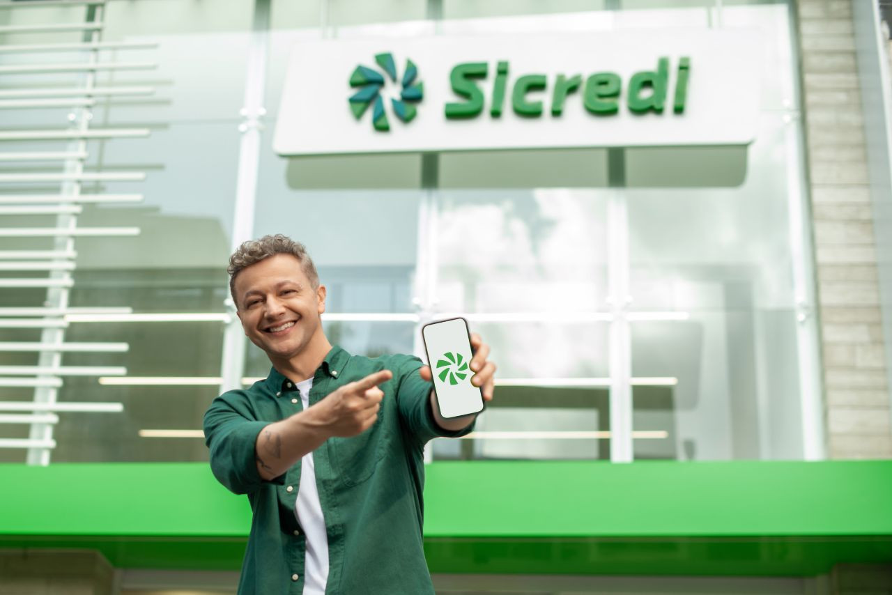 Lucas Lima sorri em frente a uma agência do Sicredi, exibindo um celular com o logo da cooperativa, em destaque na nova campanha da instituição financeira cooperativa.