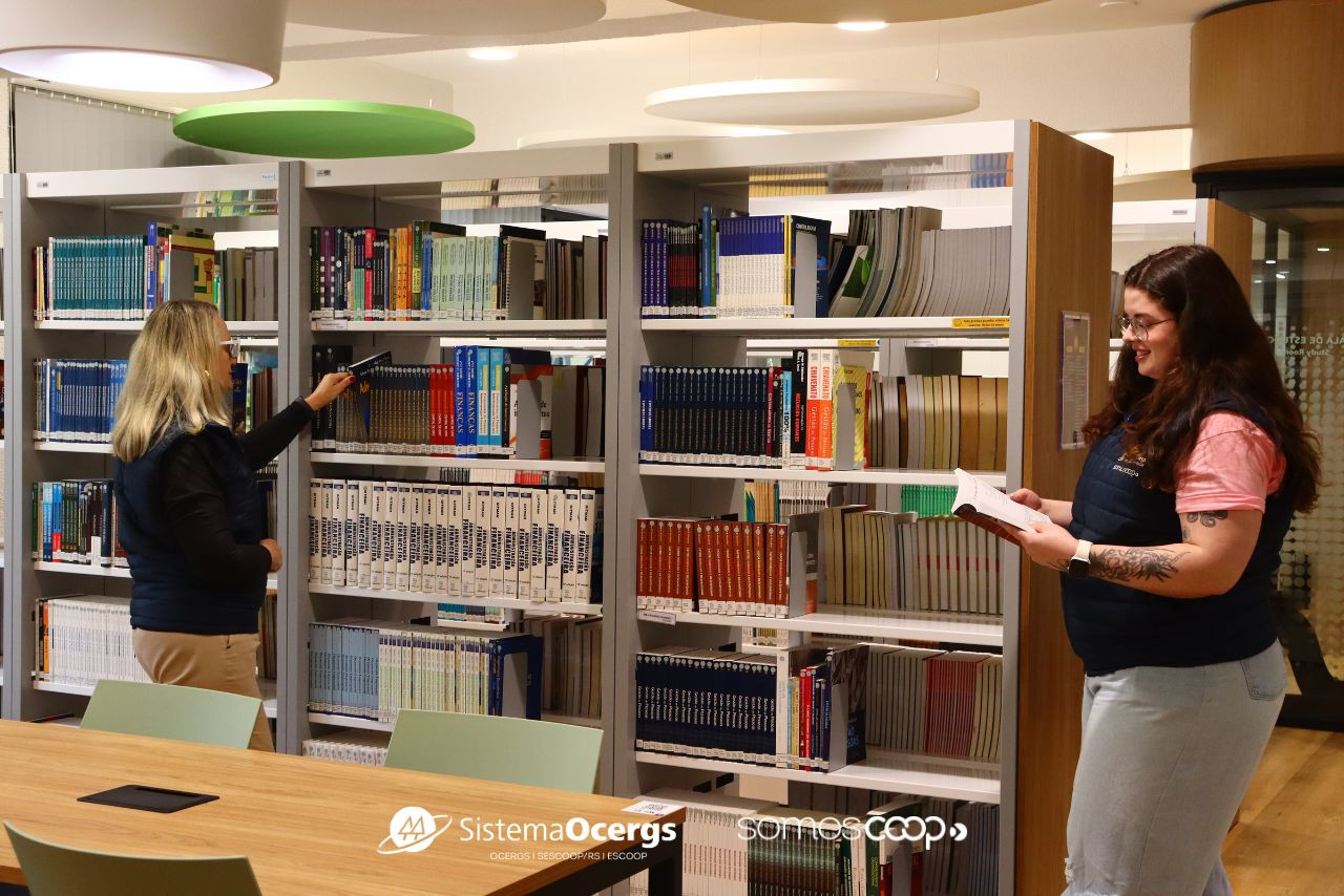 Colaboradoras do Sistema Ocergs consultam livros em estantes da Biblioteca Vergilio Frederico Perius, em ambiente moderno e acolhedor, voltado ao cooperativismo.